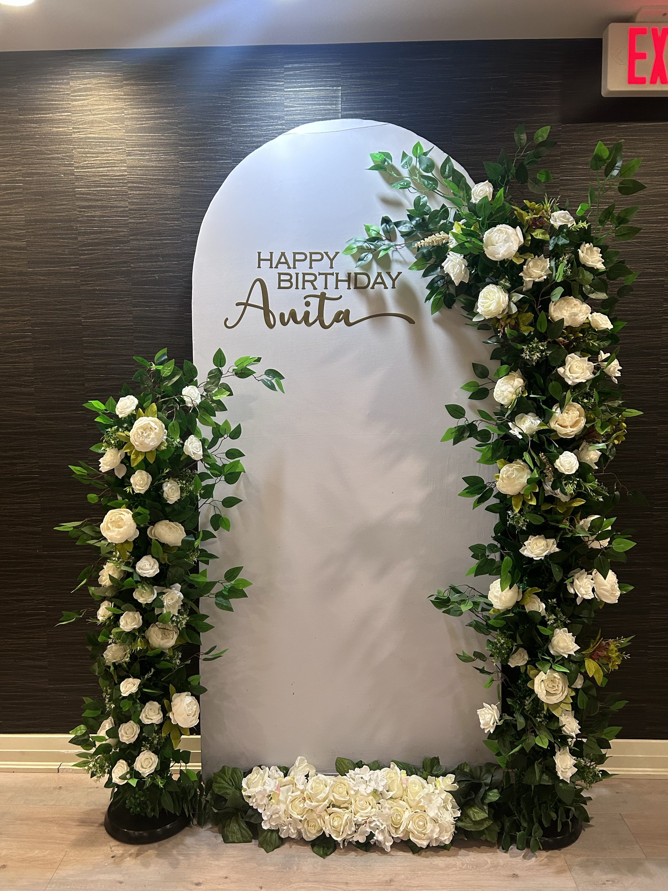A birthday arch dressed in white roses and greenery, lettered in gold